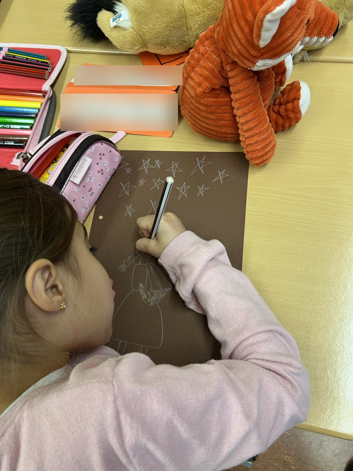 Ein Mädchen malt an seinem Schulplatz mit hellem Stift Sterne und eine Märchenfigur auf dunkles Papier