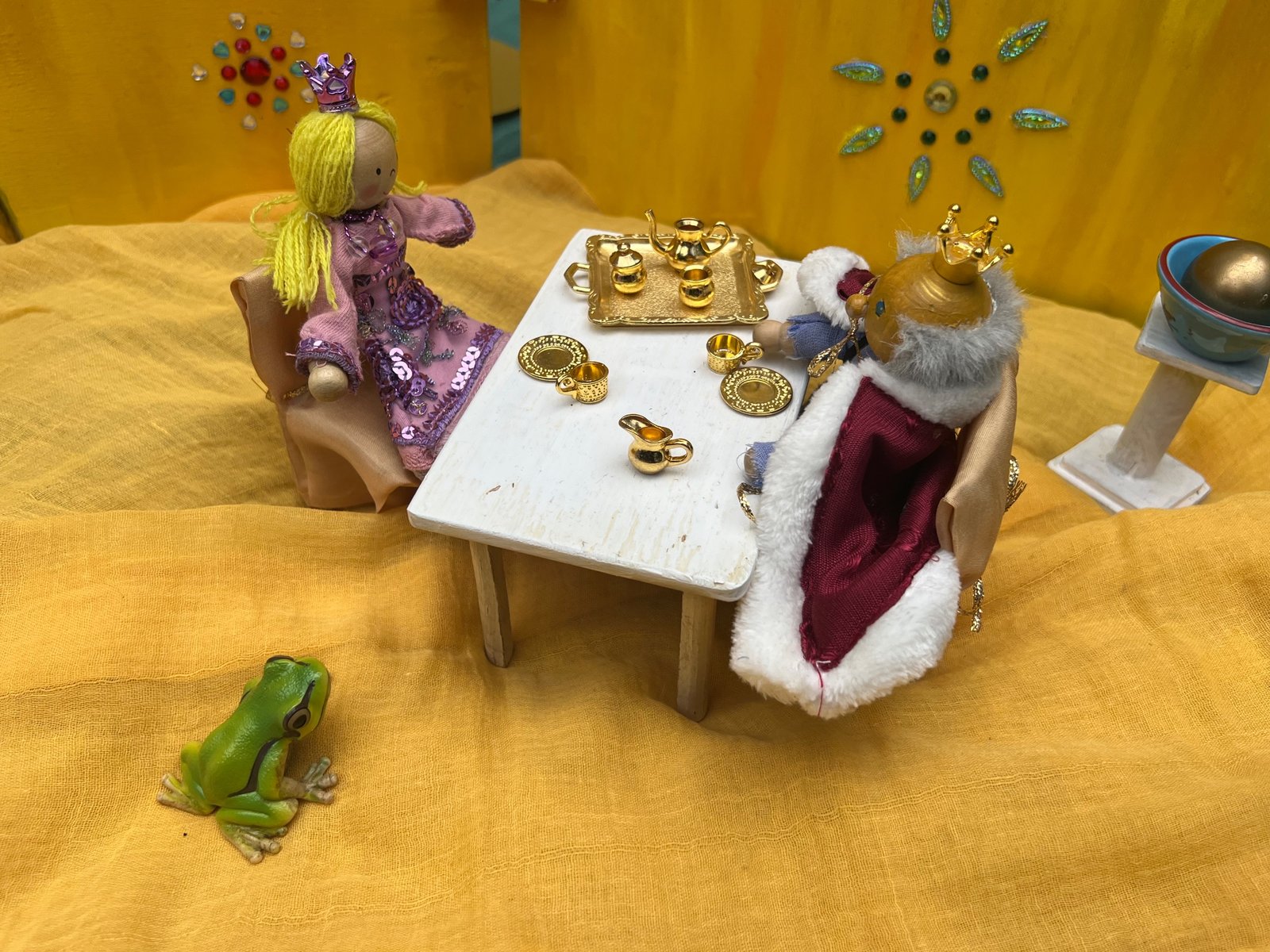 Spielszene zum Froschkönig: Prinzessin und König als Holzfiguren am gedeckten Tisch mit goldenem Geschirr, davor ein Frosch